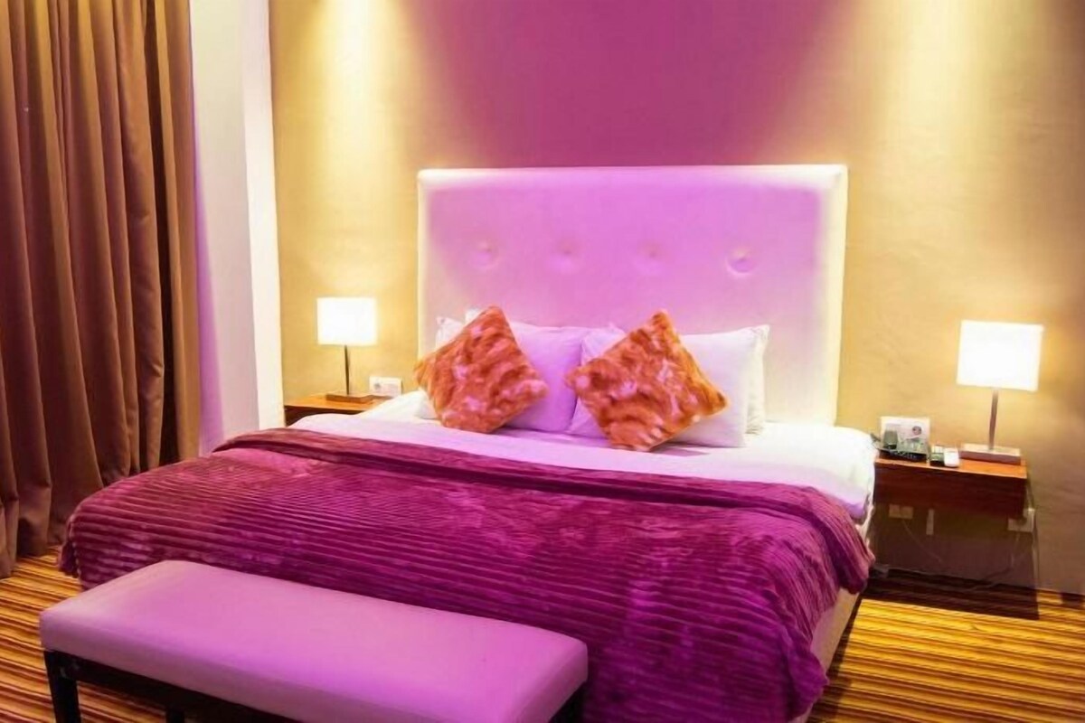Una habitación de hotel con una cama, un banco, dos mesitas de noche y cortinas.