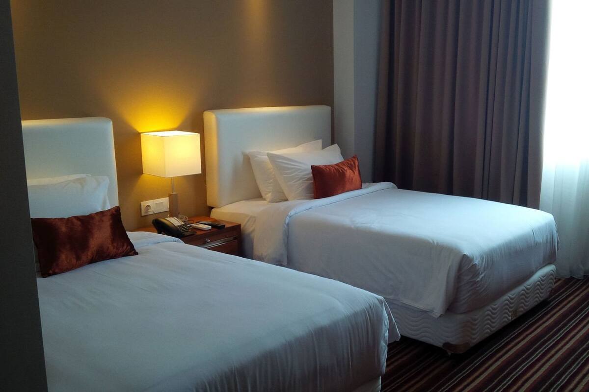 A hotel room with two beds, a bedside table with a lamp, a phone, and a window with curtains.