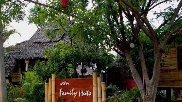 A rustic wooden hut with a thatched roof, surrounded by lush greenery and a bamboo fence. A signboard reads "Family Huts". - Family Huts (Pai)