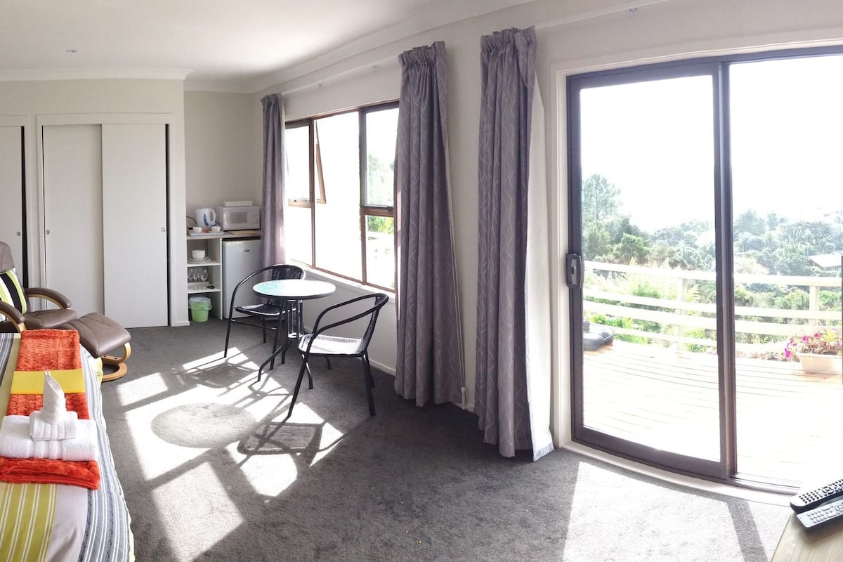 Habitación de hotel con cama, mesita de noche, lámpara y vistas al exterior a través de un ventanal grande.