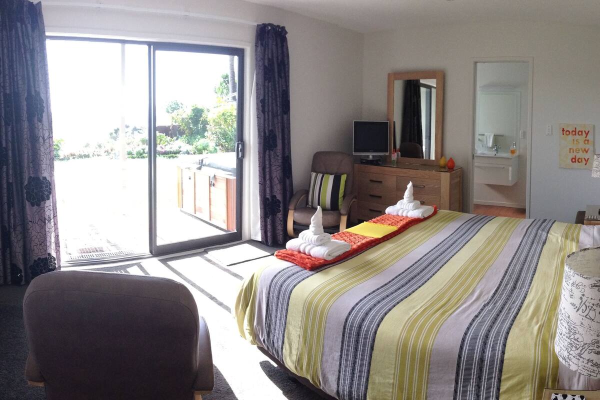 Habitación de hotel con cama, televisión, cómoda, espejo, lámpara y vistas al exterior.