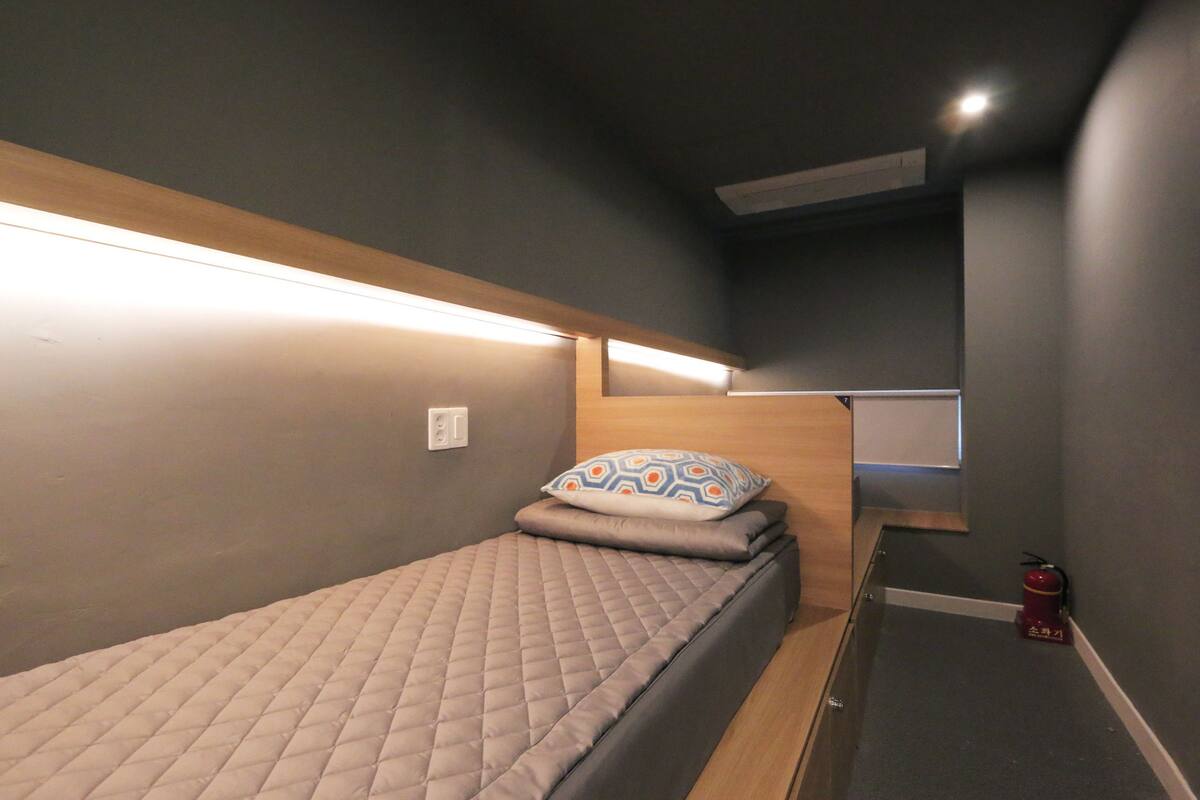 Une petite pièce, de taille modeste, comprenant un lit simple, une table de chevet et une étagère fixée au mur.