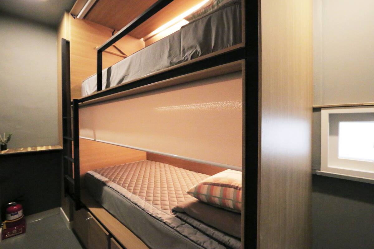 Un lit superposé avec un matelas et des oreillers, une petite table de chevet avec un récipient rouge, et une fenêtre avec des stores.