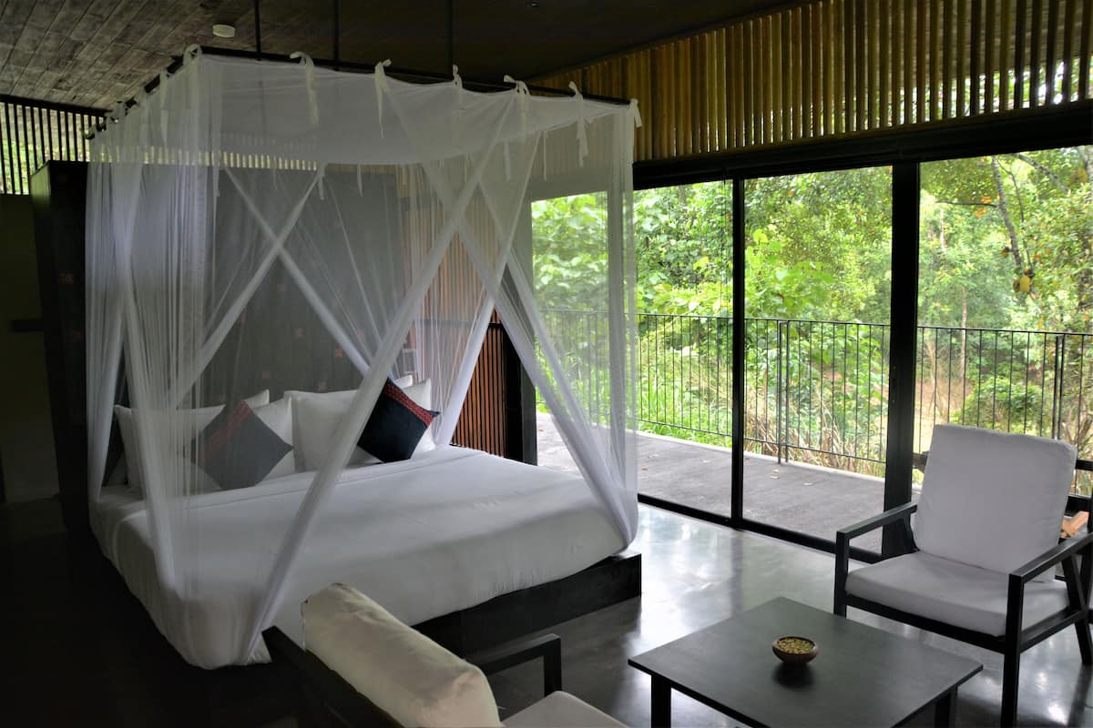 Una habitación con cama bajo un dosel, un balcón con vistas a zonas verdes y un rincón de estar con una mesa.