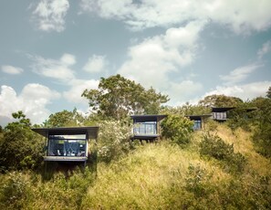 A modern, elevated structure with large windows, surrounded by lush greenery and trees, set against a partly cloudy sky.
