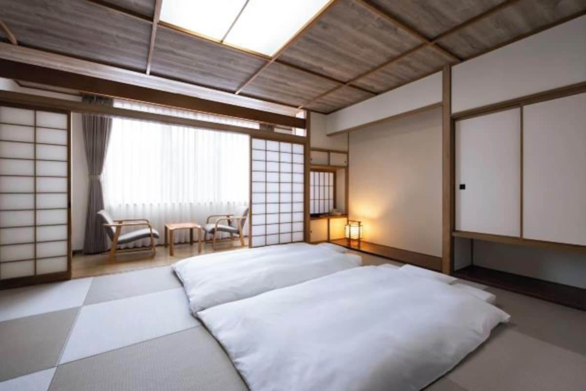 Una habitación tradicional japonesa con suelo de tatami, techo bajo, puertas correderas y una cama con ropa blanca.