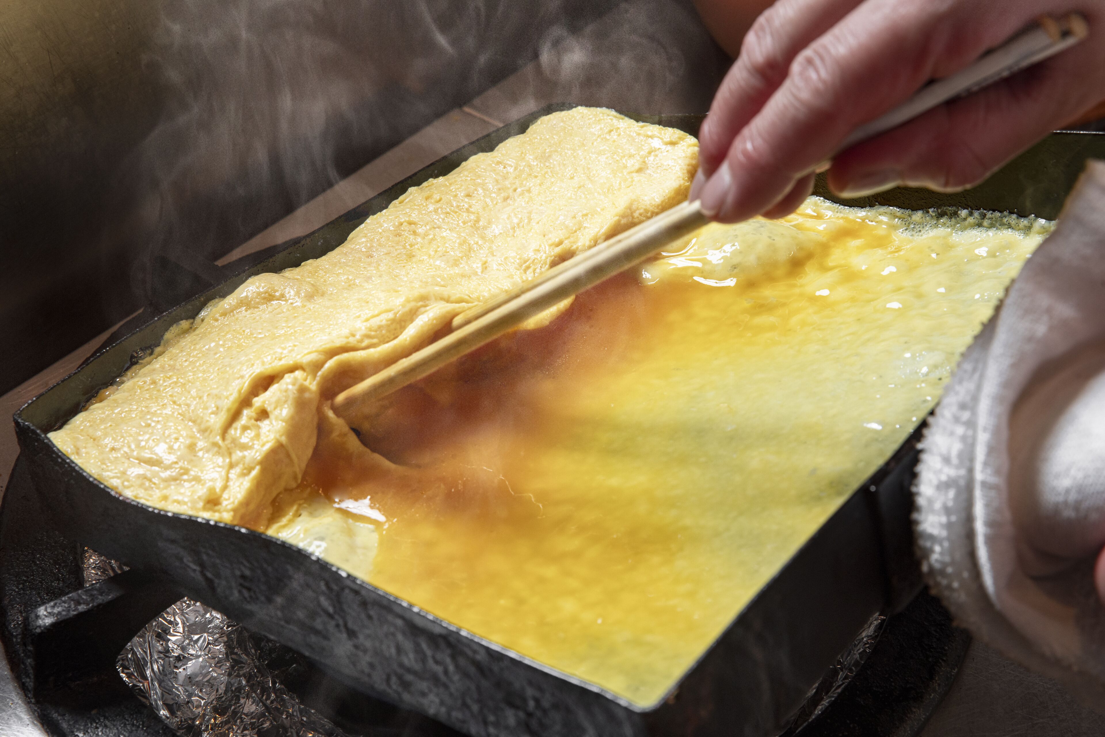 A person cooking a dish in a pan with a lid, steam rising from the pan.