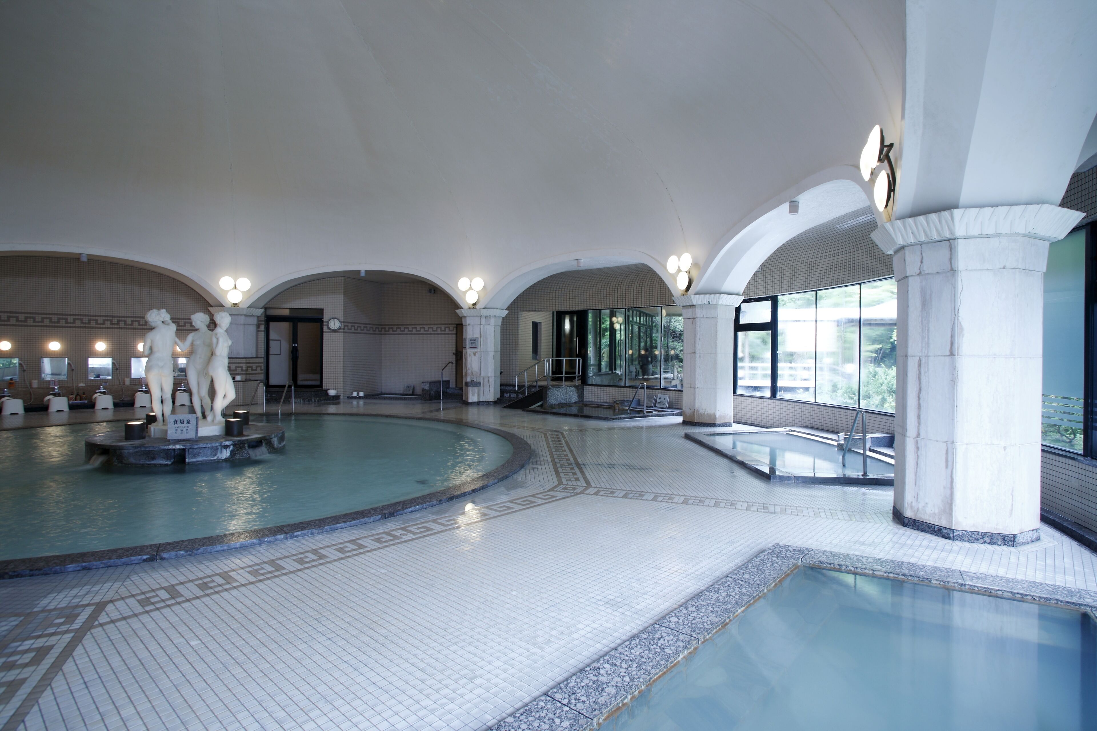 A large indoor swimming pool with a central statue, arched ceilings, and a tiled floor.