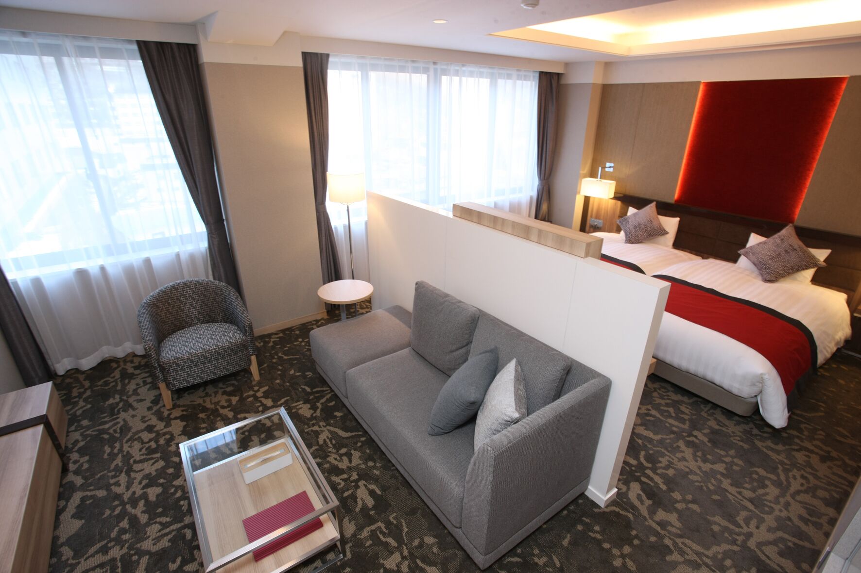A hotel room with a grey sofa, a small round table, a chair, a bed with a red headboard, and a large window with curtains.