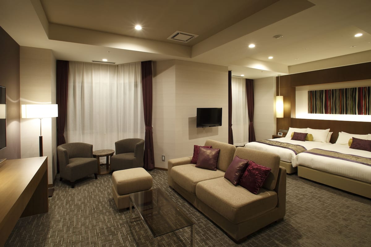A hotel room with a sofa, armchair, a small table, a bed, and a TV mounted on the wall.