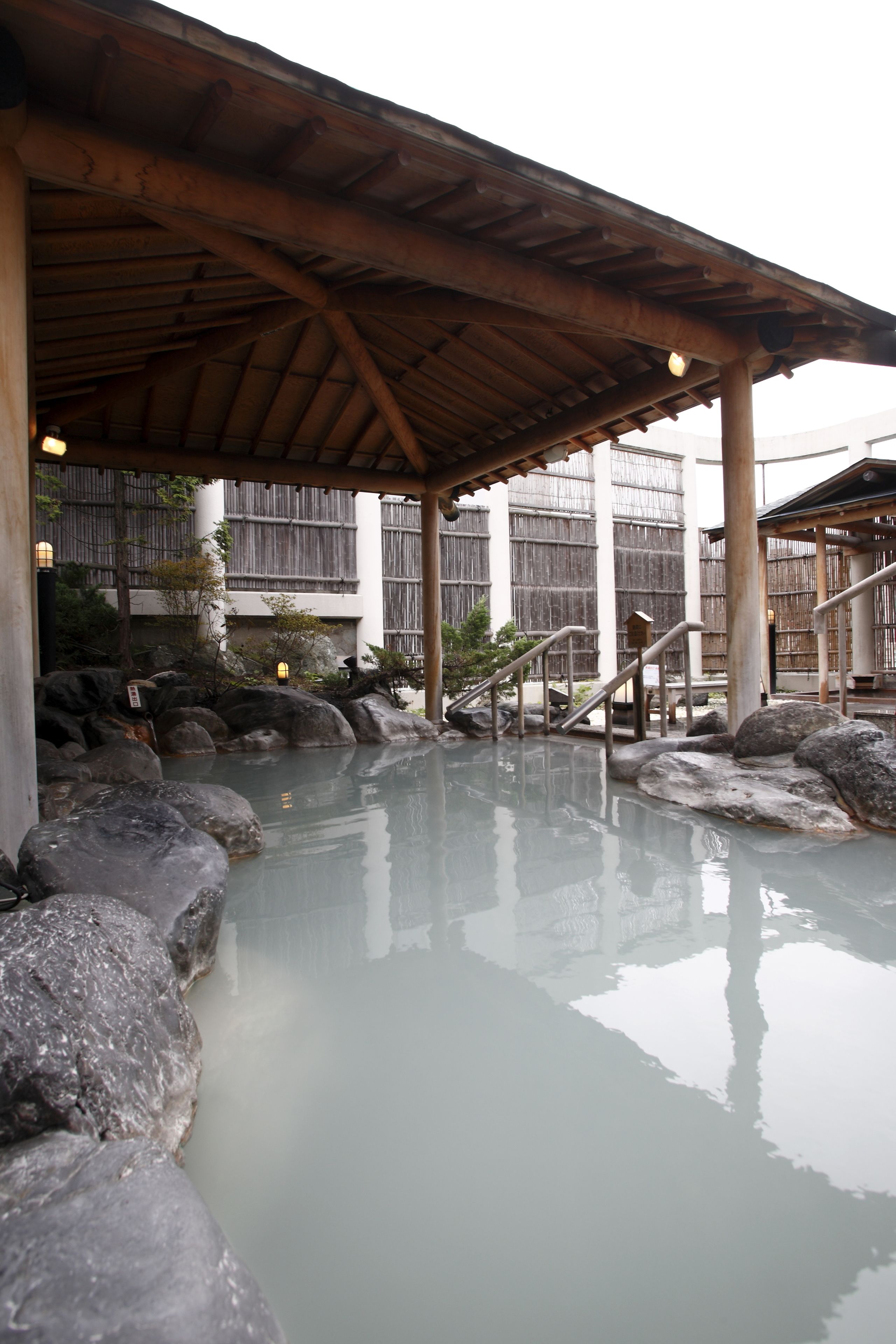 A traditional Japanese onsen with a wooden roof, stone steps, and a bamboo fence in the background.