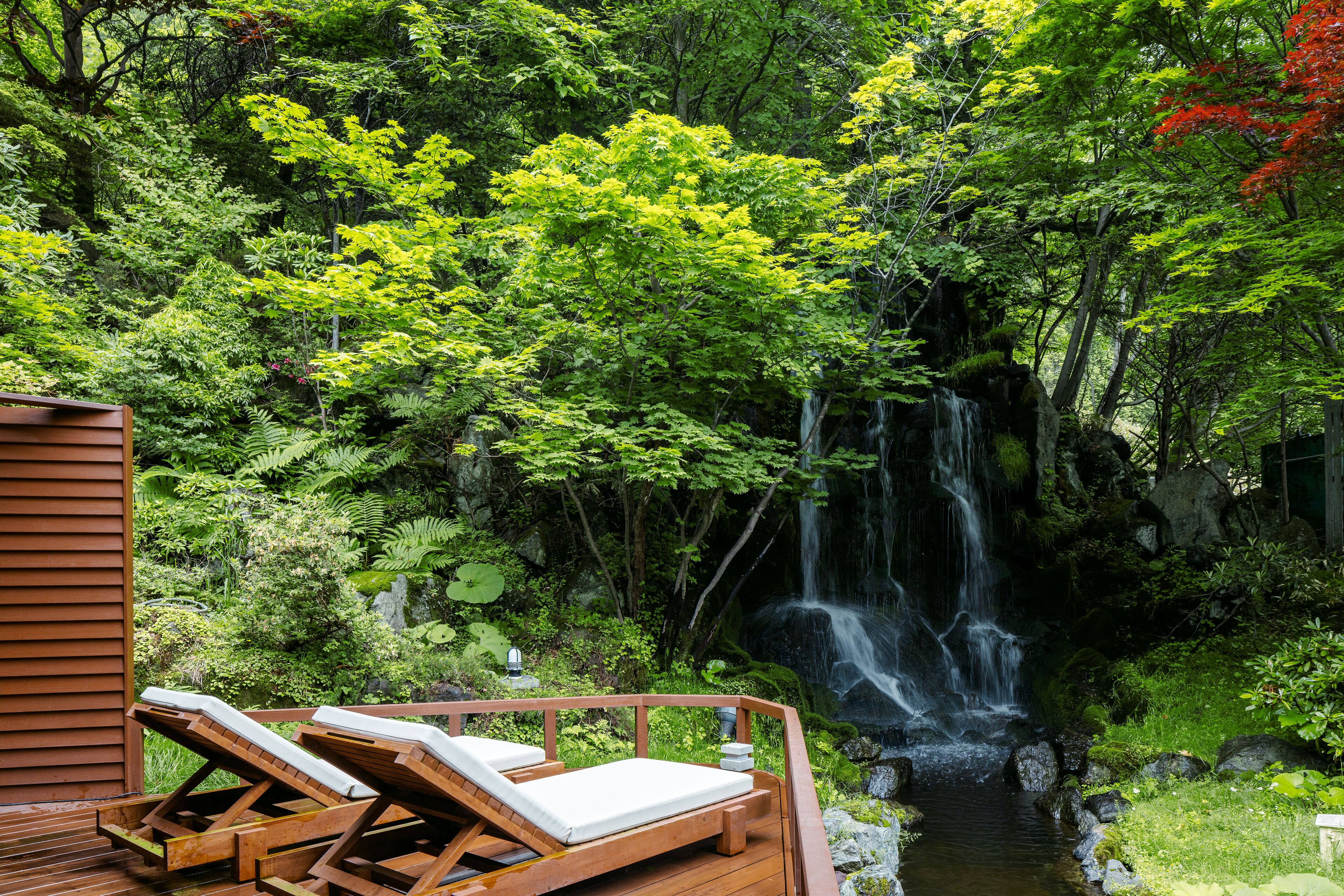 A wooden deck with two lounge chairs facing a waterfall in a lush forest.