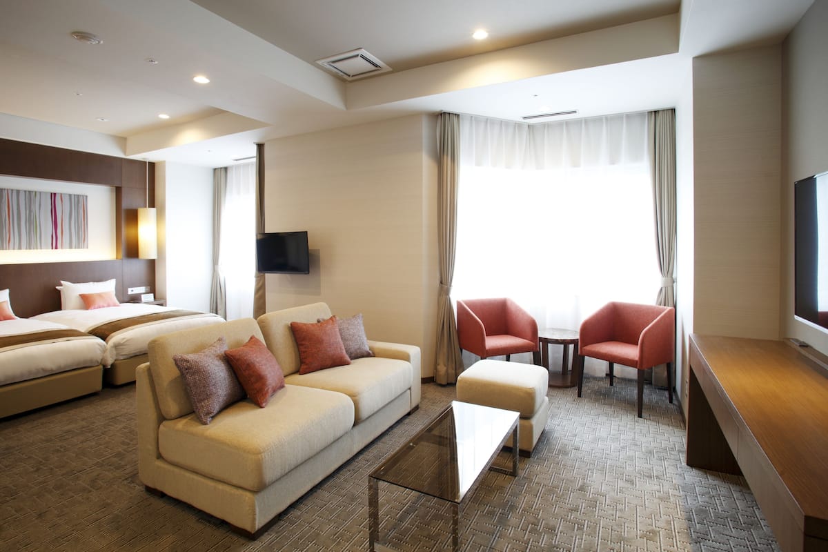 A hotel room with a sofa, two single beds, a TV, a desk, and a window with curtains.