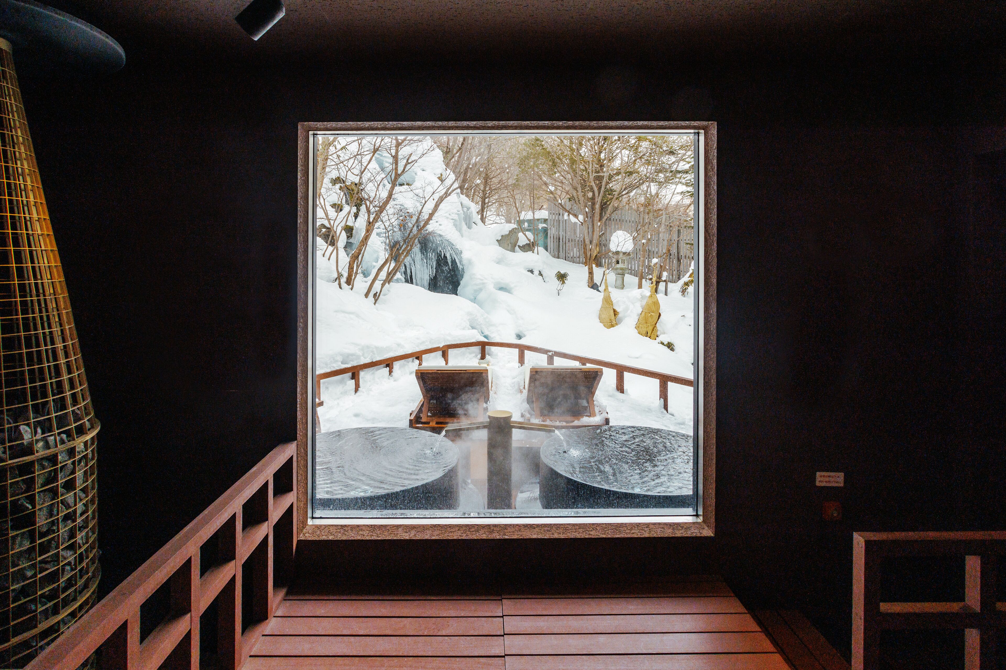 A view through a window of a wooden deck with two chairs and a small table, overlooking a snowy landscape with trees and a wooden bridge.