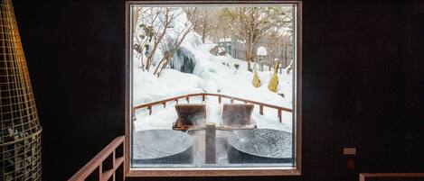 A view through a window of a wooden deck with two chairs and a small table, overlooking a snowy landscape with trees and a wooden bridge.