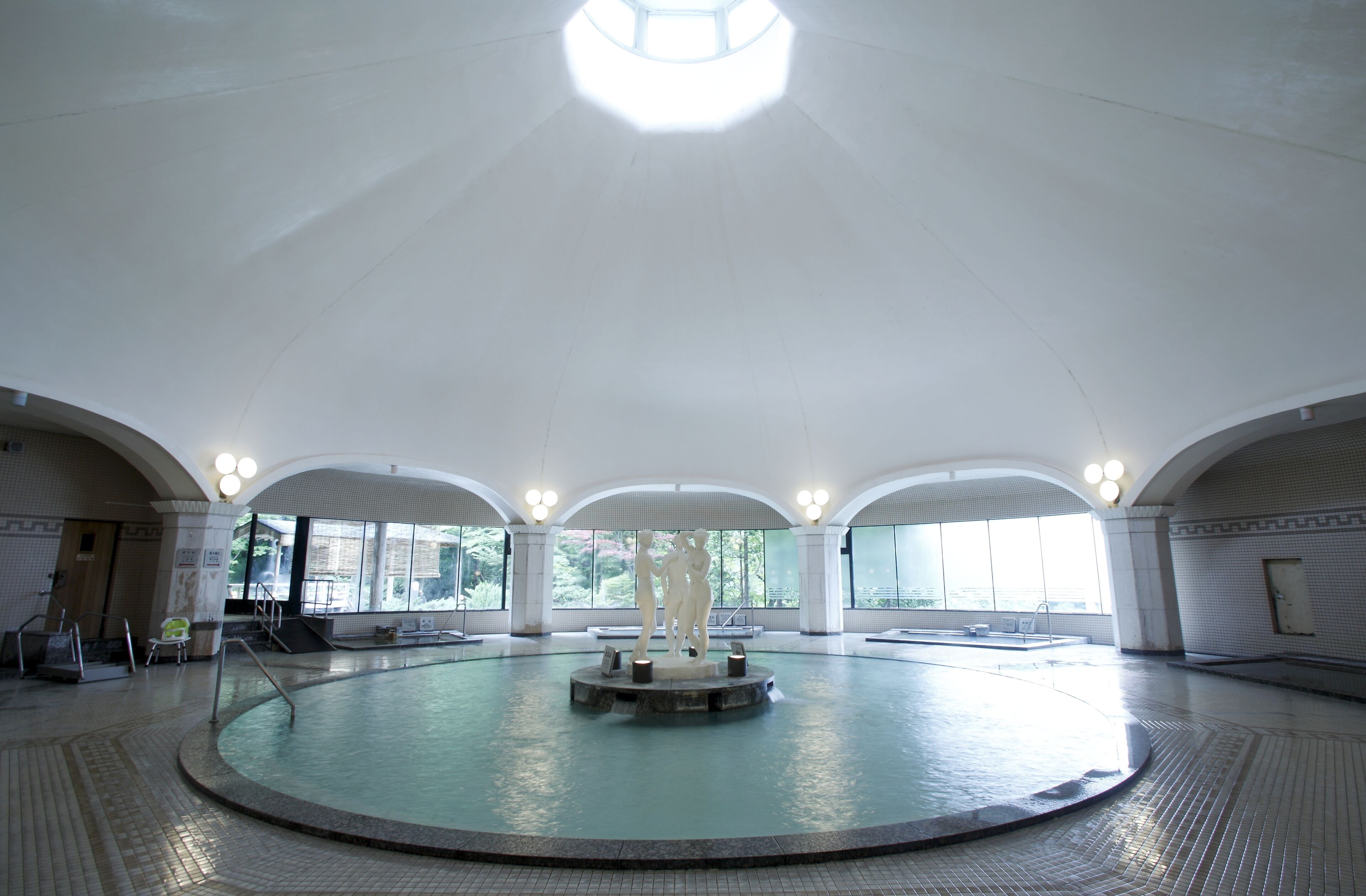 A large indoor swimming pool with a central sculpture, arched ceilings, and large windows.