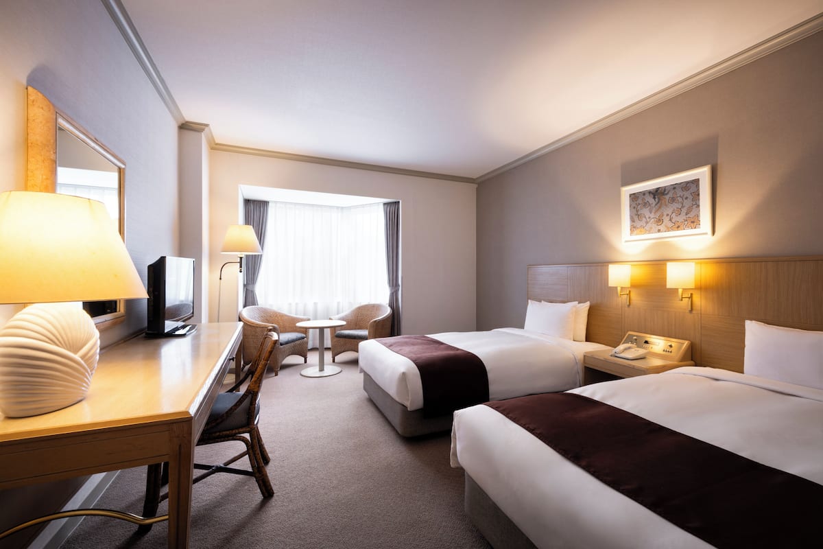 A hotel room with two beds, a desk with a lamp, a chair, a small table, a TV, and a window with curtains.