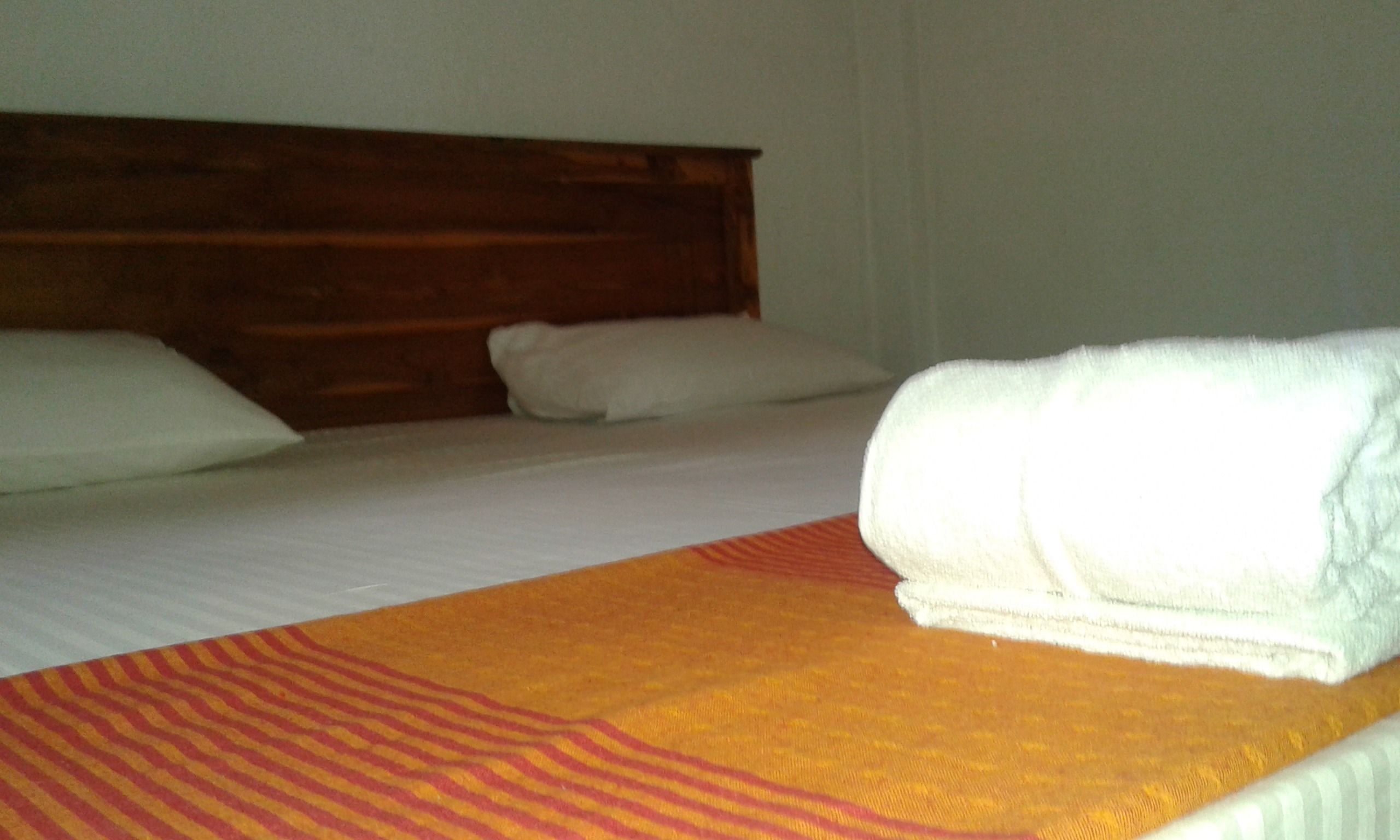 Habitación de hotel con cabecera de madera, cama con sábanas blancas y una toalla blanca enrollada sobre un corredor de cama naranja a rayas.