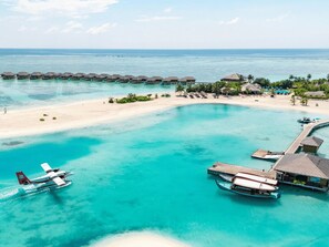 Vista aérea de un complejo turístico con bungalós sobre el agua, una playa de arena y aguas turquesas cristalinas.