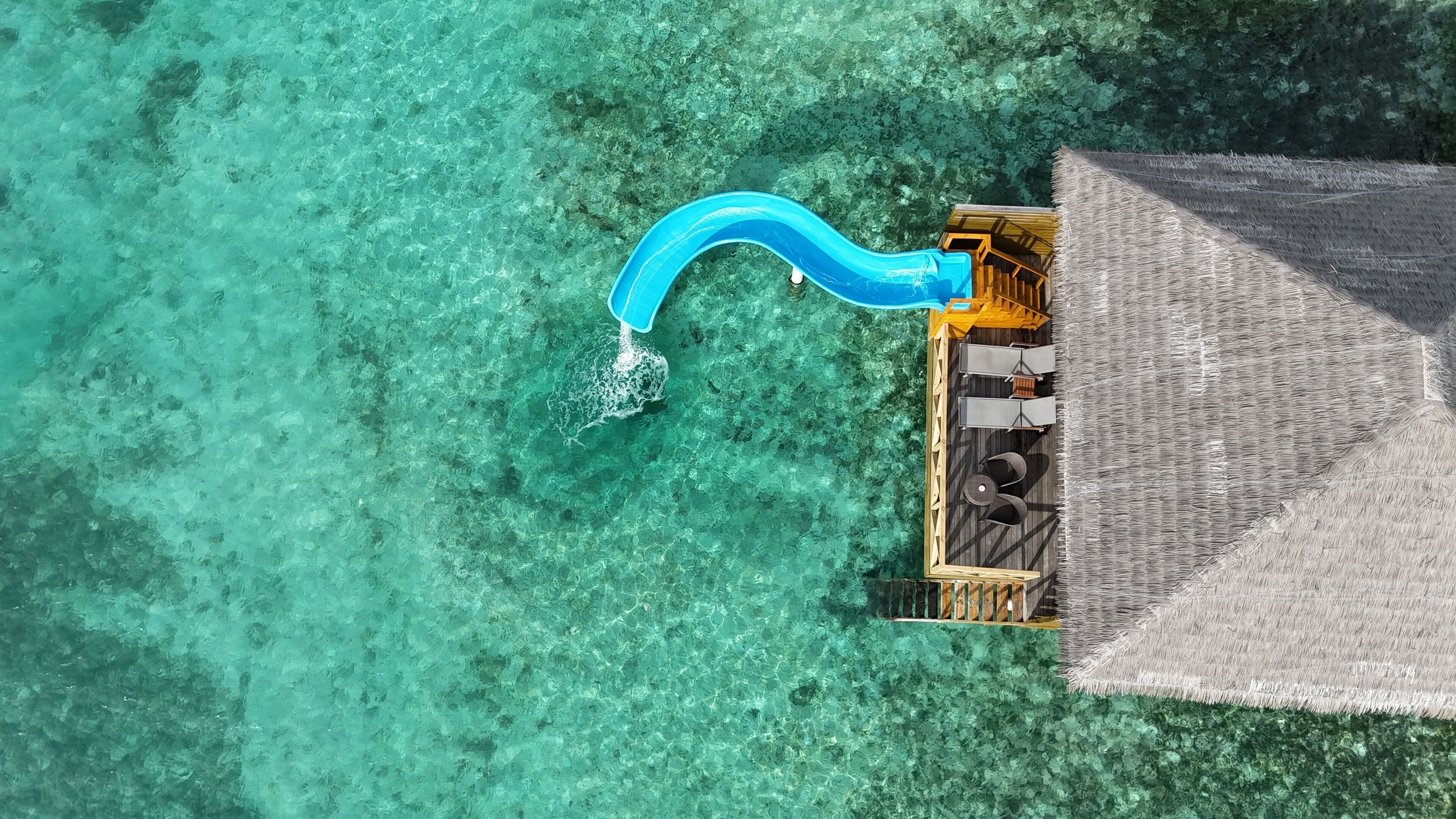 An aerial view of a resort with a clear turquoise sea, a thatched-roof structure, and a blue water slide.