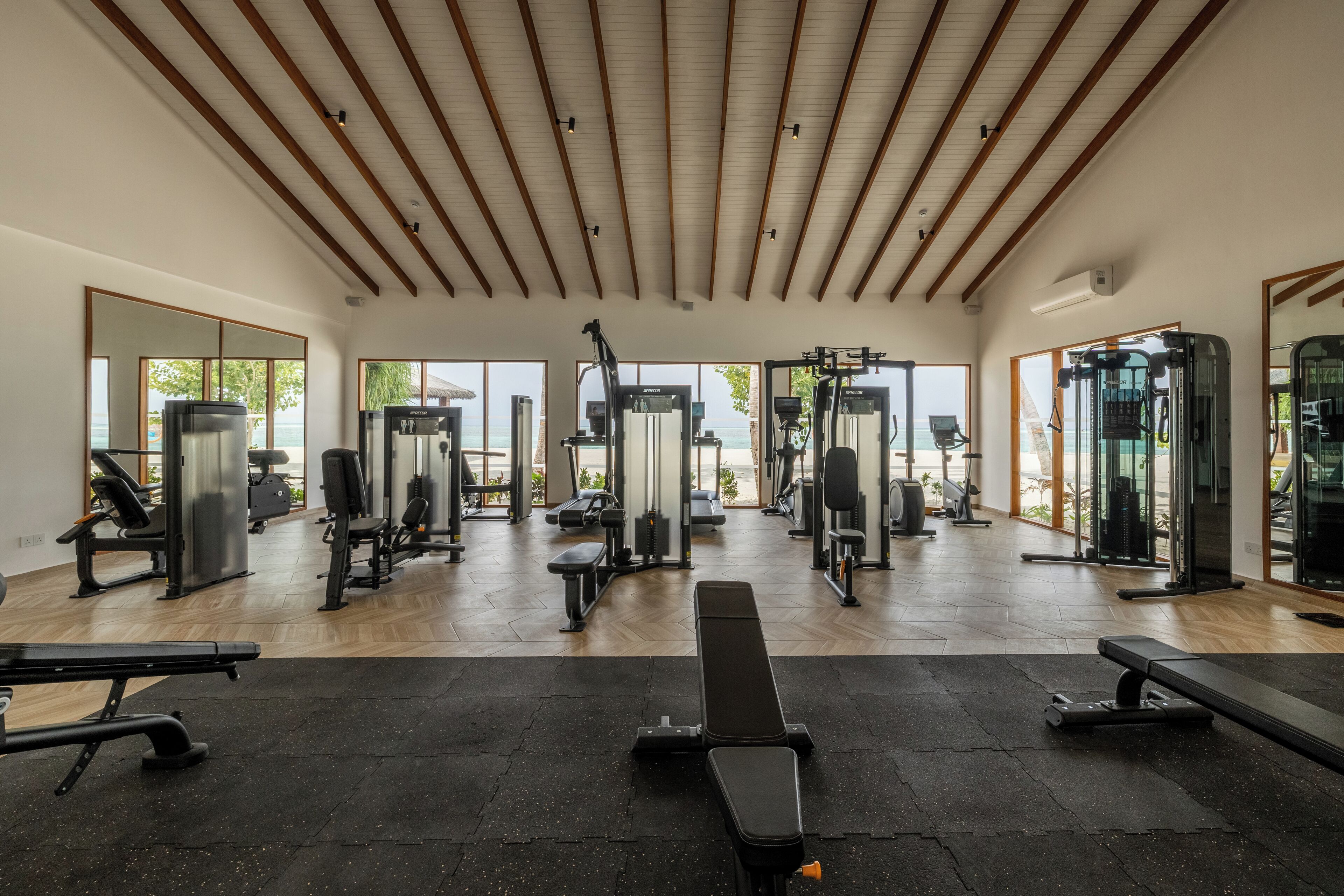 A spacious gym with various exercise equipment, large windows, and a wooden ceiling.