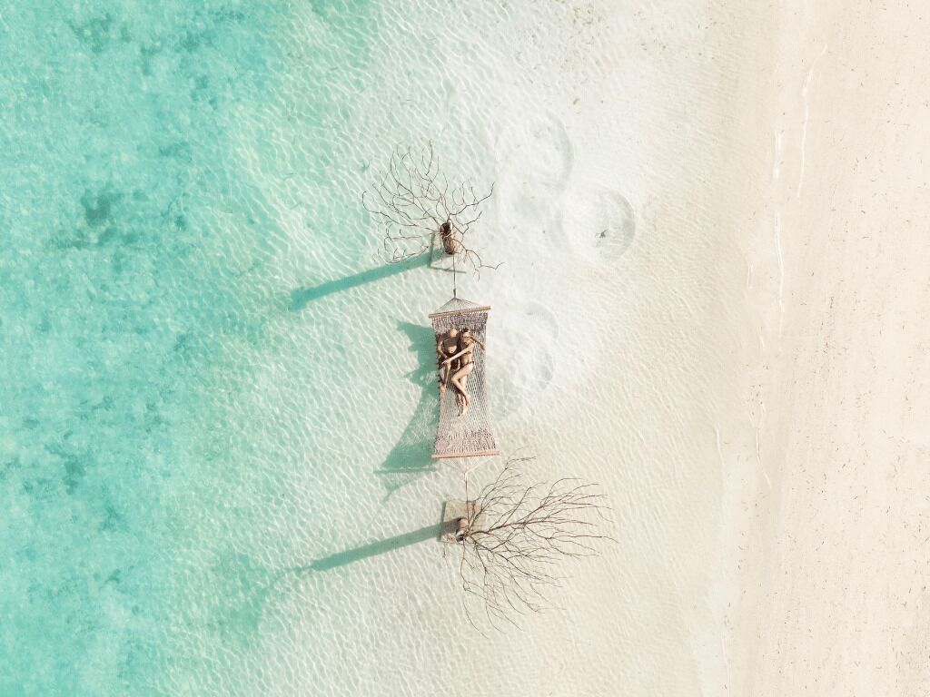 An aerial view of a beach with clear turquoise water, a sandy shore, and a few structures including a birdcage and a small tree.