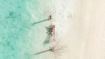 An aerial view of a beach with clear turquoise water, a sandy shore, and a few structures including a birdcage and a small tree.