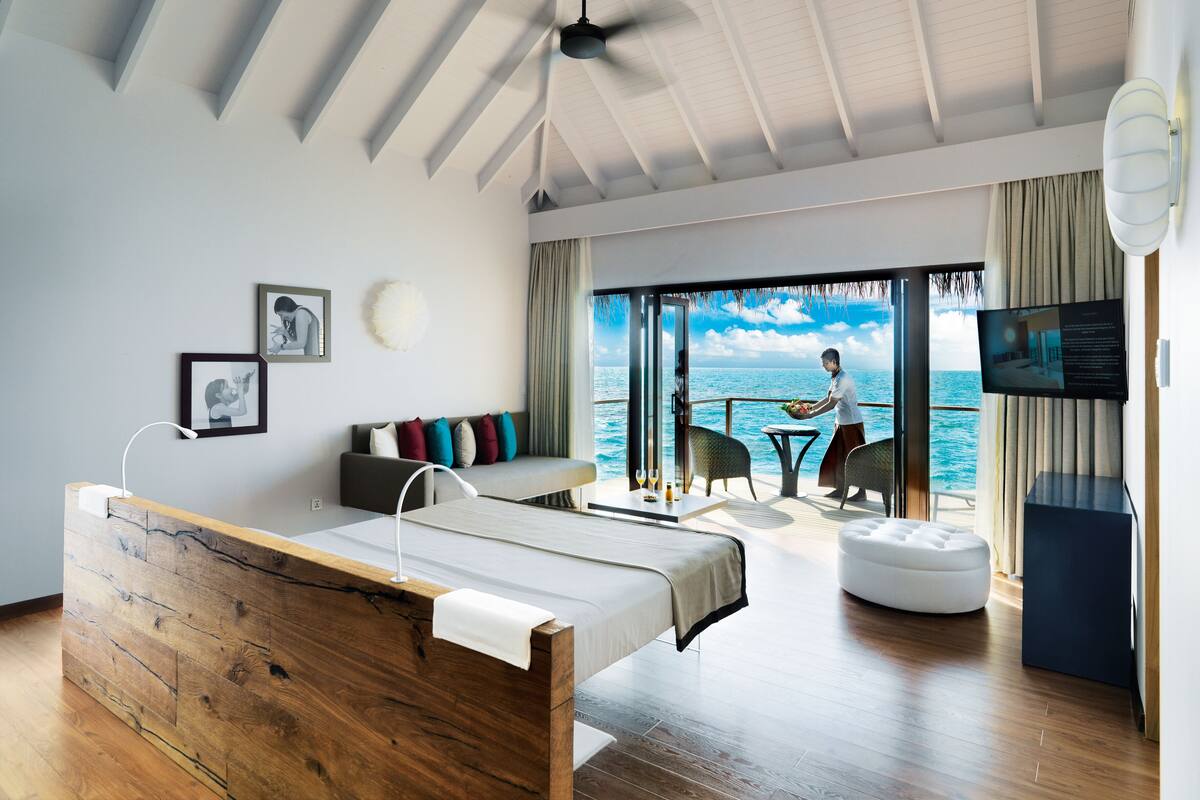 Une chambre d’hôtel moderne avec un lit en bois, un canapé agrémenté de coussins colorés, un coin repas avec une table et des chaises, et une grande fenêtre offrant une vue sur la mer.