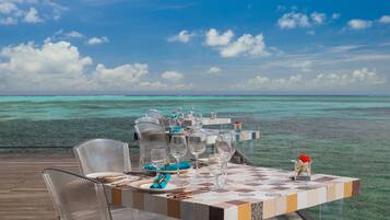Une table à manger, avec ses chaises, ses verres et ses couverts, est installée sur une terrasse offrant une vue sur l’océan.