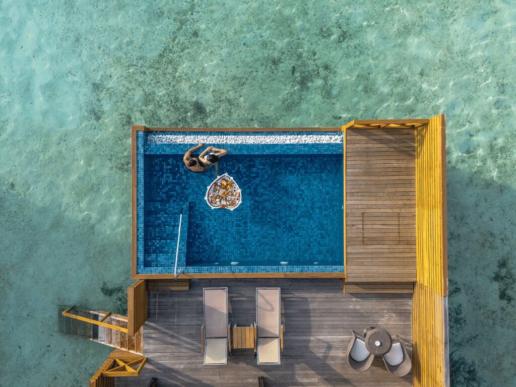 Vista aérea de un área de piscina con muelle de madera, sillas de descanso y un pequeño bote.