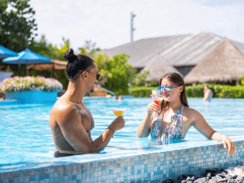 Una pareja en una piscina, con una bebida en la mano, rodeada de follaje tropical y construcciones de techo de paja.