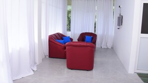 Une pièce avec deux canapés rouges, des rideaux blancs, un téléviseur à écran plat fixé au mur et un sol en béton.