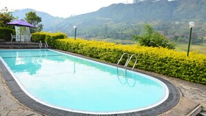 Une piscine bordée de pierres, une petite table et des chaises placées sous un parasol violet, et une vue sur des collines verdoyantes en arrière-plan.