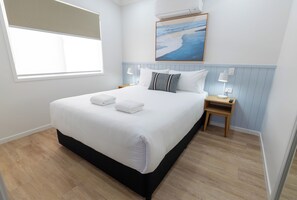 Een opgemaakt bed met witte lakens en twee handdoeken, een nachtkastje met een lamp, een wandairconditioner en een schilderij van een strand.