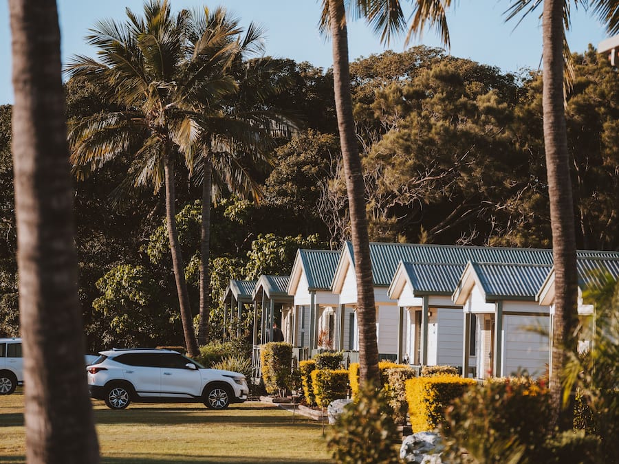A row of white cabins with green roofs, surrounded by palm trees and greenery.
