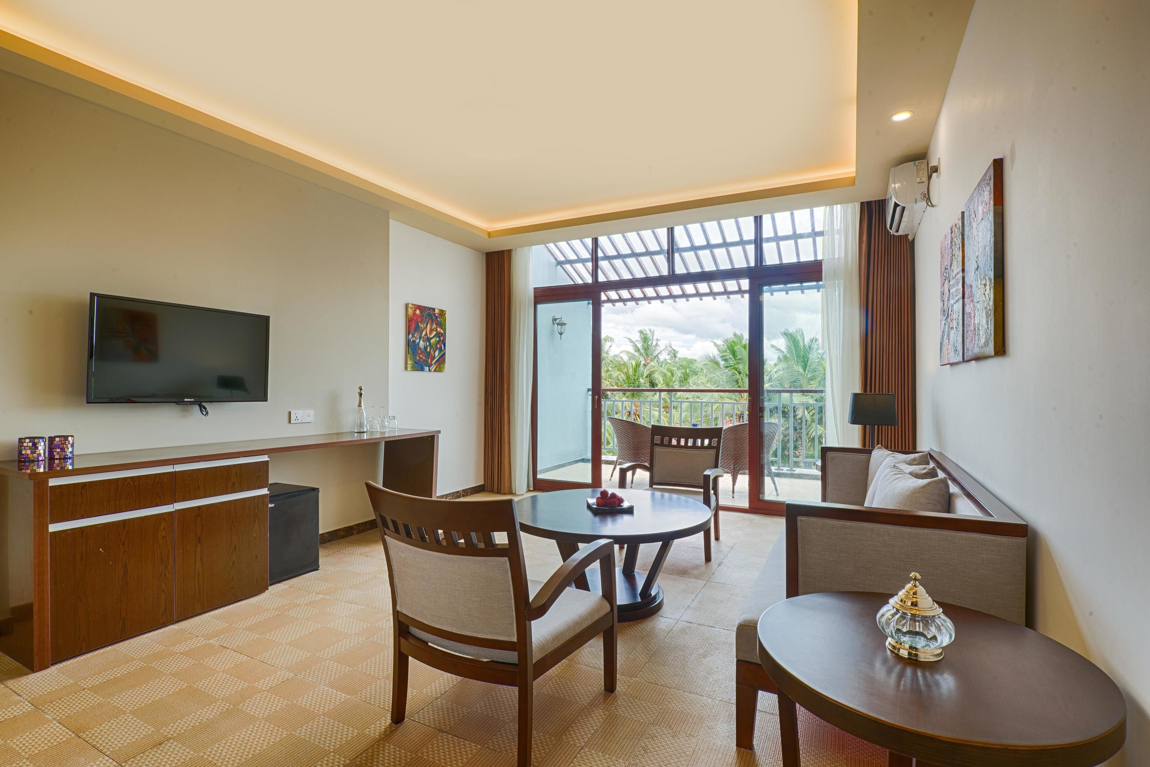 A modern hotel room with a flat-screen TV, a dining area with a round table and chairs, a sofa, and a balcony with a view of palm trees.