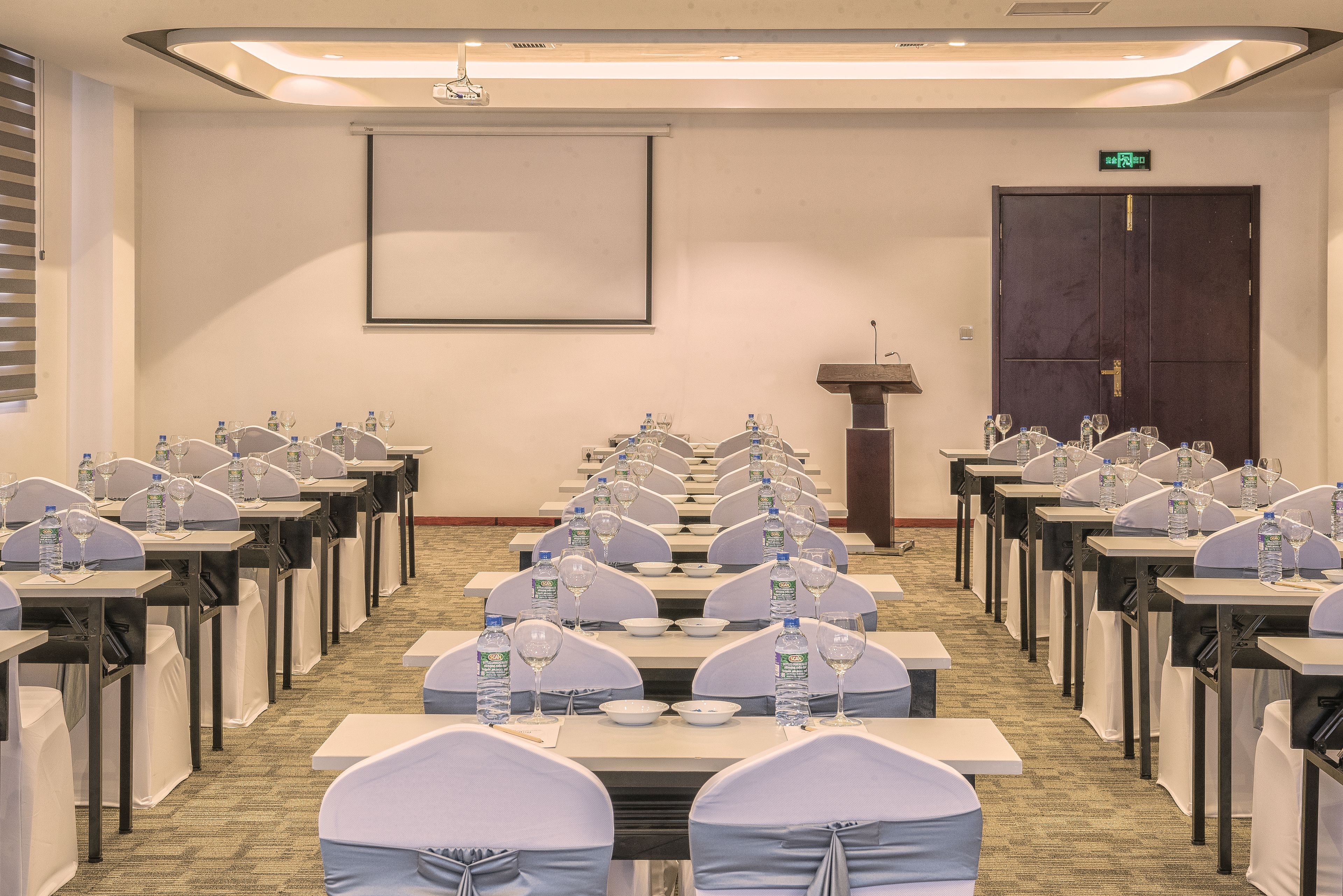 Une salle de conférence avec des rangées de tables, chacune étant équipée de bouteilles d’eau, de verres à vin et de chaises recouvertes d’un tissu blanc.