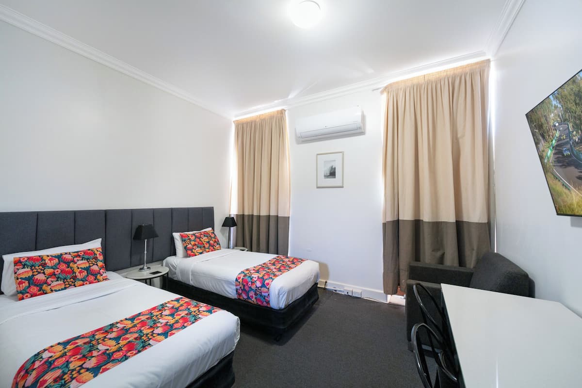 A hotel room with two beds, a desk, a wheelchair, and a painting on the wall.