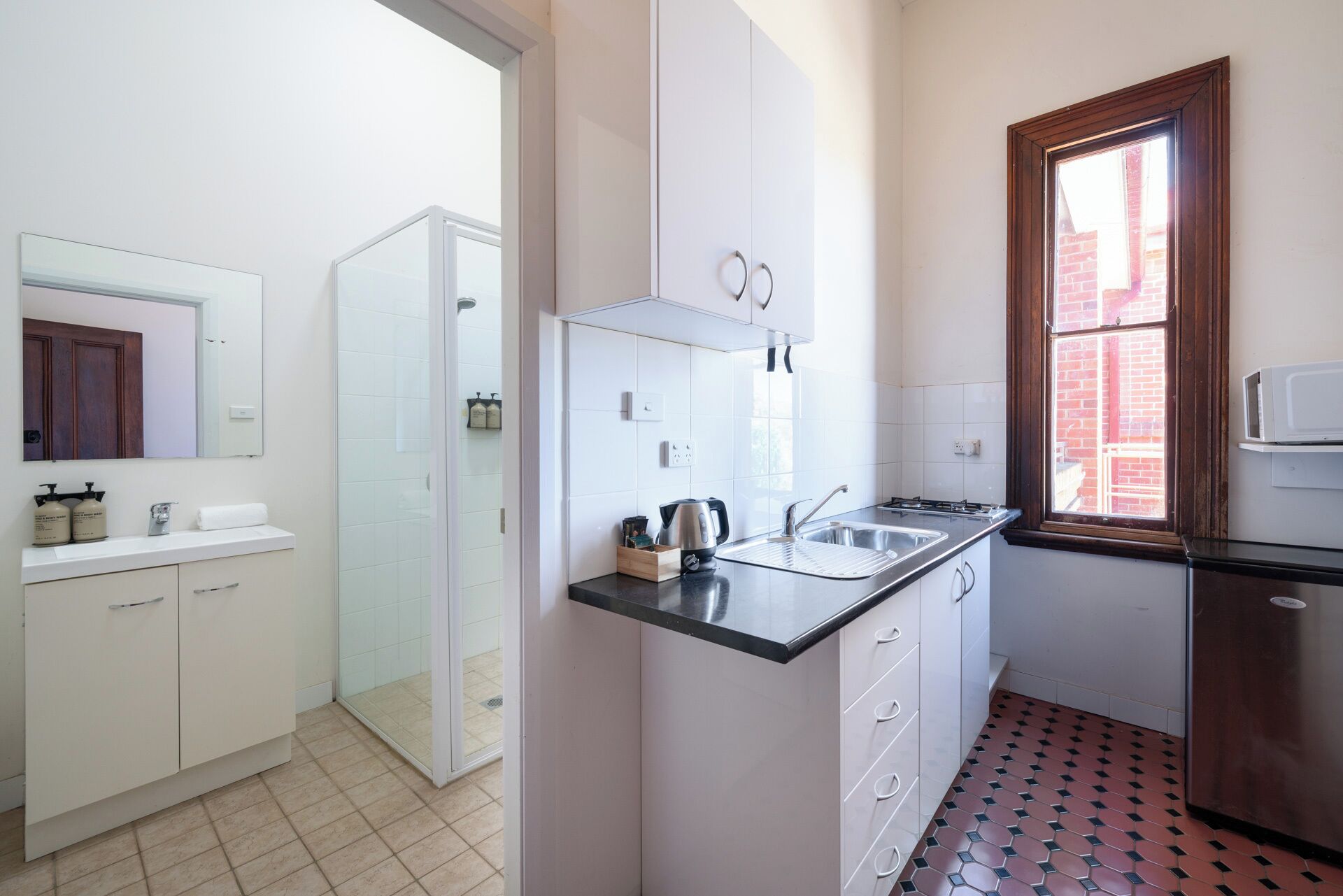 Cocina compacta con armarios blancos, encimera negra y ventana con vista a un edificio de ladrillo.