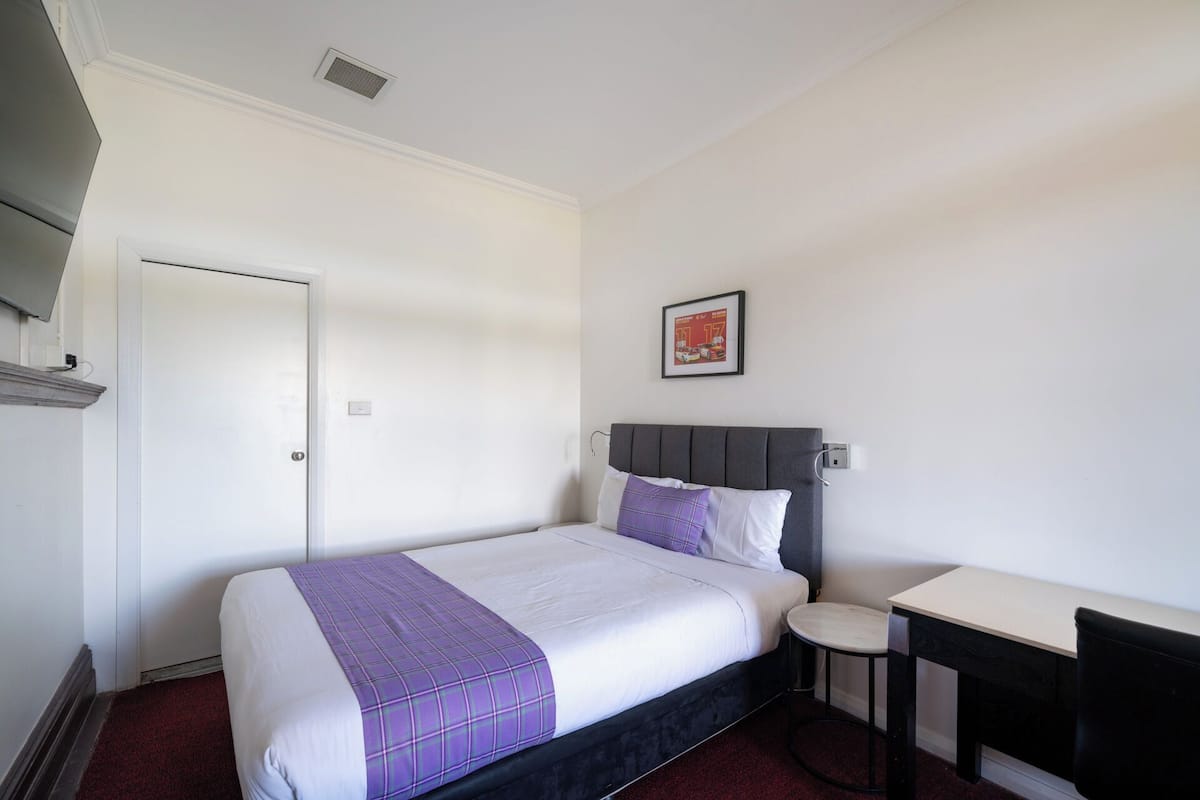 A hotel room with a bed, a small desk, a chair, a door, and a wall-mounted picture.