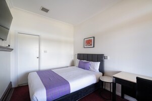 Habitación de hotel con cama, un escritorio pequeño, una silla, una puerta y un cuadro colgado en la pared.