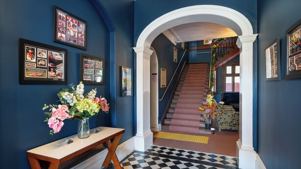 A hallway with a checkered floor, framed pictures on the walls, a wooden table with a vase of flowers, and a staircase with a railing. - The Hallows Accommodation (South Bathurst)