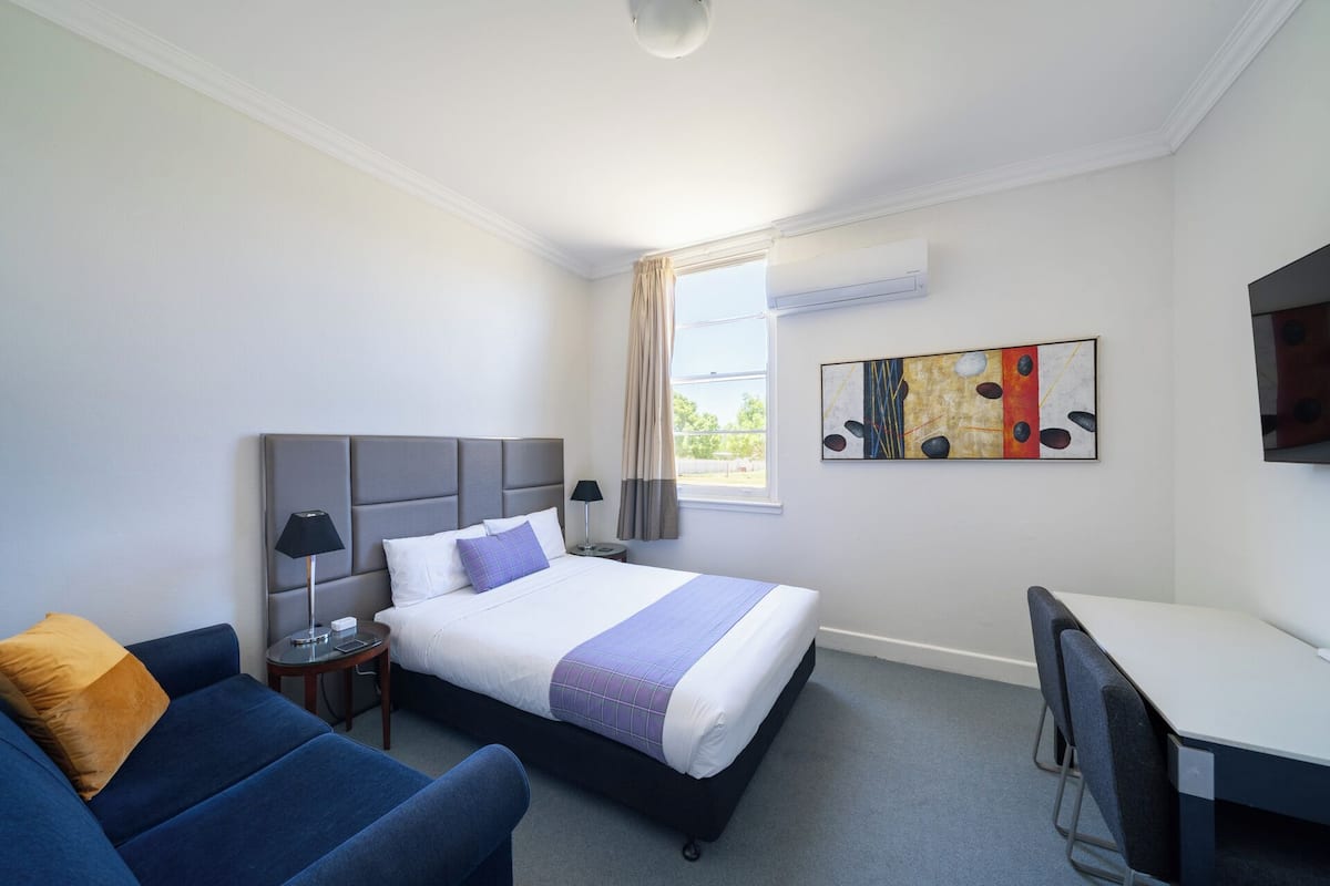 A hotel room with a bed, a desk, a chair, a sofa, and a painting on the wall.