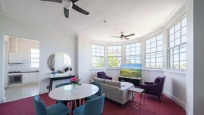 A modern living room with a round table, blue chairs, a sofa, and a TV. There are large windows with a view of greenery. - The Hallows Accommodation (South Bathurst)