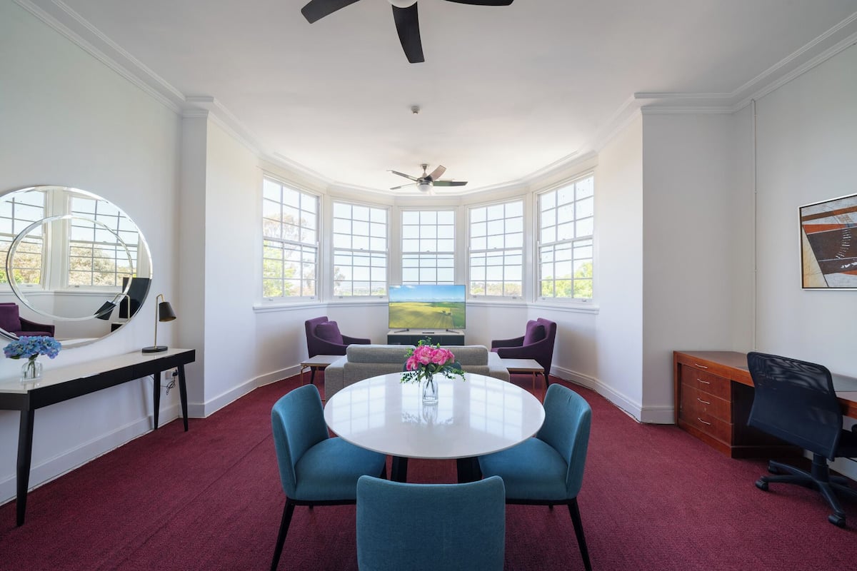 A room with a round table, four chairs, a TV, a ceiling fan, and large windows.