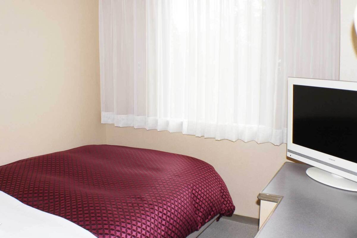 A hotel room with a bed, a TV, and a window with sheer curtains.