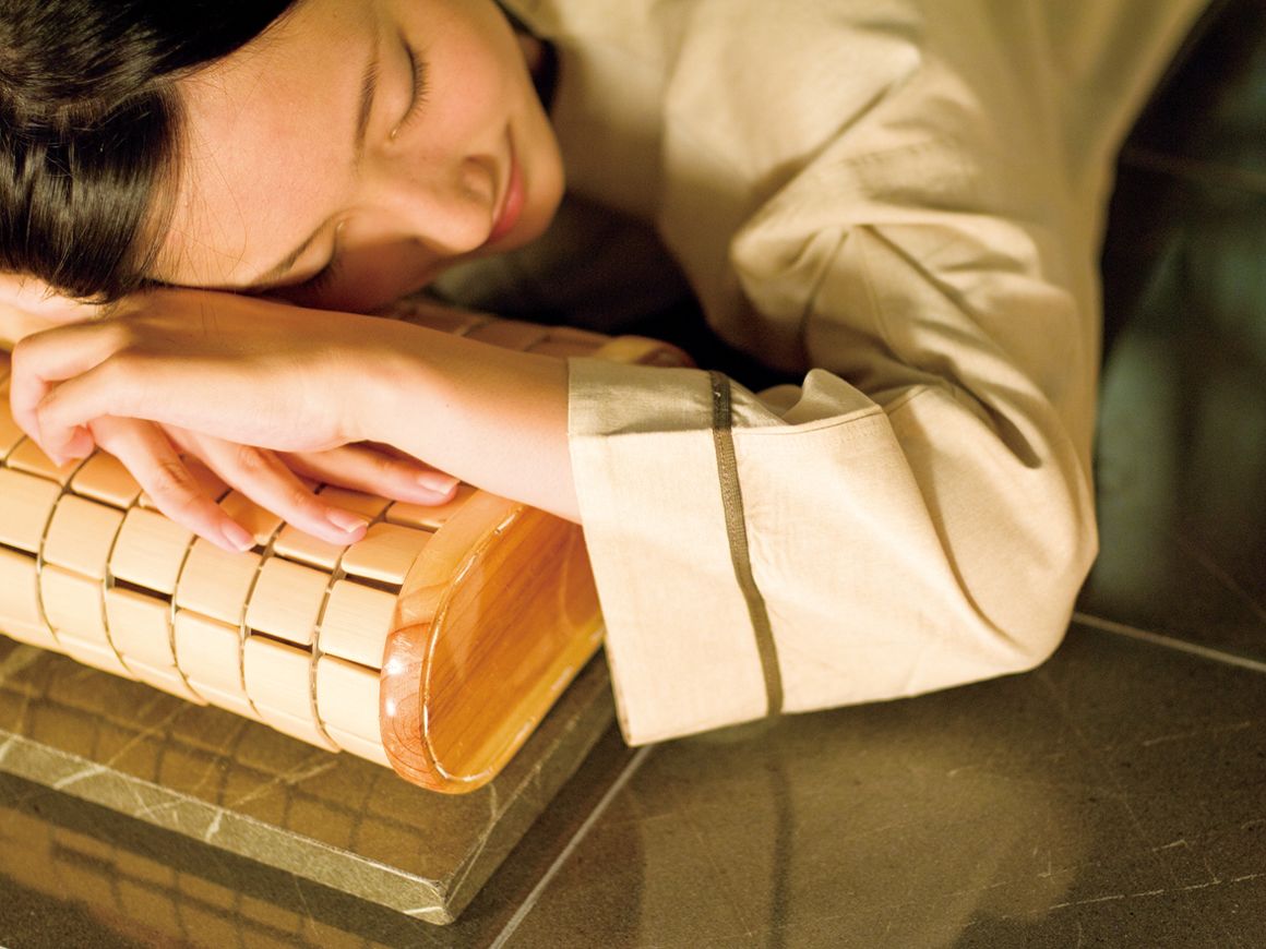 A person resting their head on a wooden object, possibly a massage tool, in a room with a tiled floor.