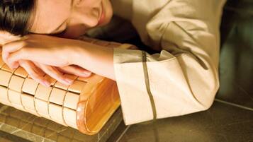 A person resting their head on a wooden object, possibly a massage tool, in a room with a tiled floor.