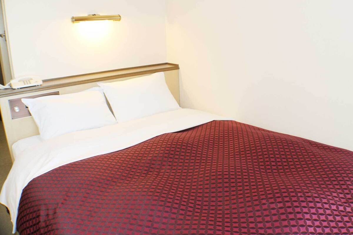 A hotel room with a bed, white bedding, a red patterned bedspread, and a bedside telephone.