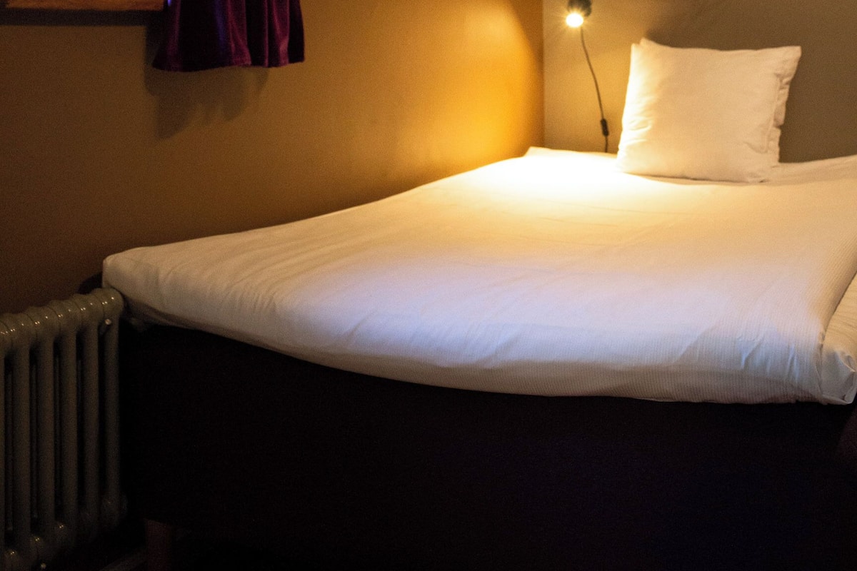 Una camera da letto con un letto, una finestra con tende, uno scaffale a parete e un termosifone.